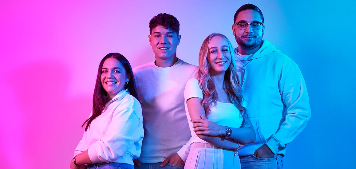 Ein Foto, auf dem vier junge Menschen erkennbar sind. Die Personen lächeln selbstbewusst in die Kamera. Im Hintergrund ist eine helle Farbstimmung erkennbar.